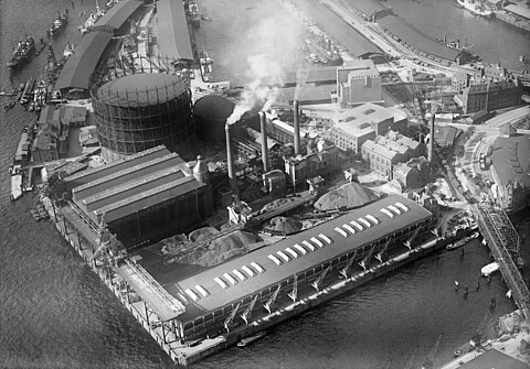 Schwarzweiße Luftaufnahme des Grasbrooks im Hamburger Hafen mit Fruchtschuppen C im Vordergrund und Gaswerk mit Gasometer im Hintergrund, um 1930