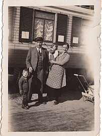 A man holding his son on the hand standing next to his wife who has a child in her arms. Both are in front of a house