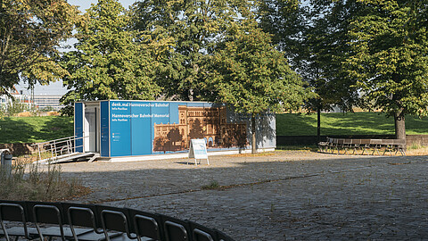 Container mit einer Rollstuhlrampe davor, beklebt mit dem Schrift und einem historischen Bild des Hannoverschen Bahnhofs, im Hintergrund Bäume, im Vordergrund ein Platz und Sitzgelegenheiten