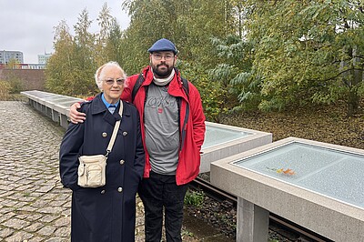 An elderly woman and a man stand in front of a row of name plaques. The man has his arm around the older woman's shoulders. Behind the name plaques are railway tracks.