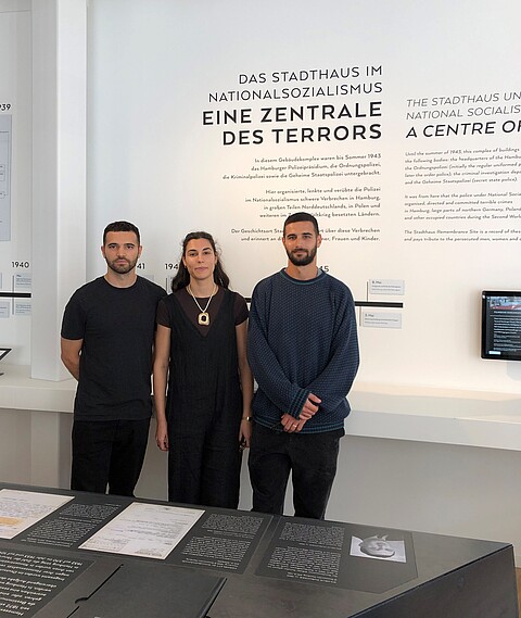 Besuch der Enkelkinder im Geschichtsort Stadthaus. Drei junge Personen, zwei Männer und eine Frau, stehen in der Ausstellung am Geschichtsort Stadthaus. Sie lächeln in die Kamera.